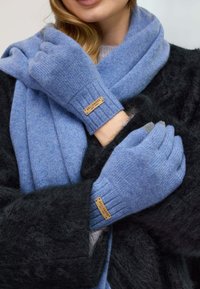 Gants tricotés bleus en matière douce, dotés de poignets côtelés et d'une petite étiquette marron. Une écharpe noire texturée est également visible.