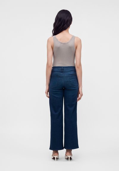 Femme aux longs cheveux foncés portant un haut beige sans manches, un jean large bleu foncé et des chaussures blanches à talons, debout de dos.
