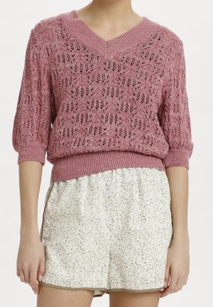 Femme portant un pull rose en maille à col en V avec des manches trois-quarts et un short blanc taille haute à motif floral.