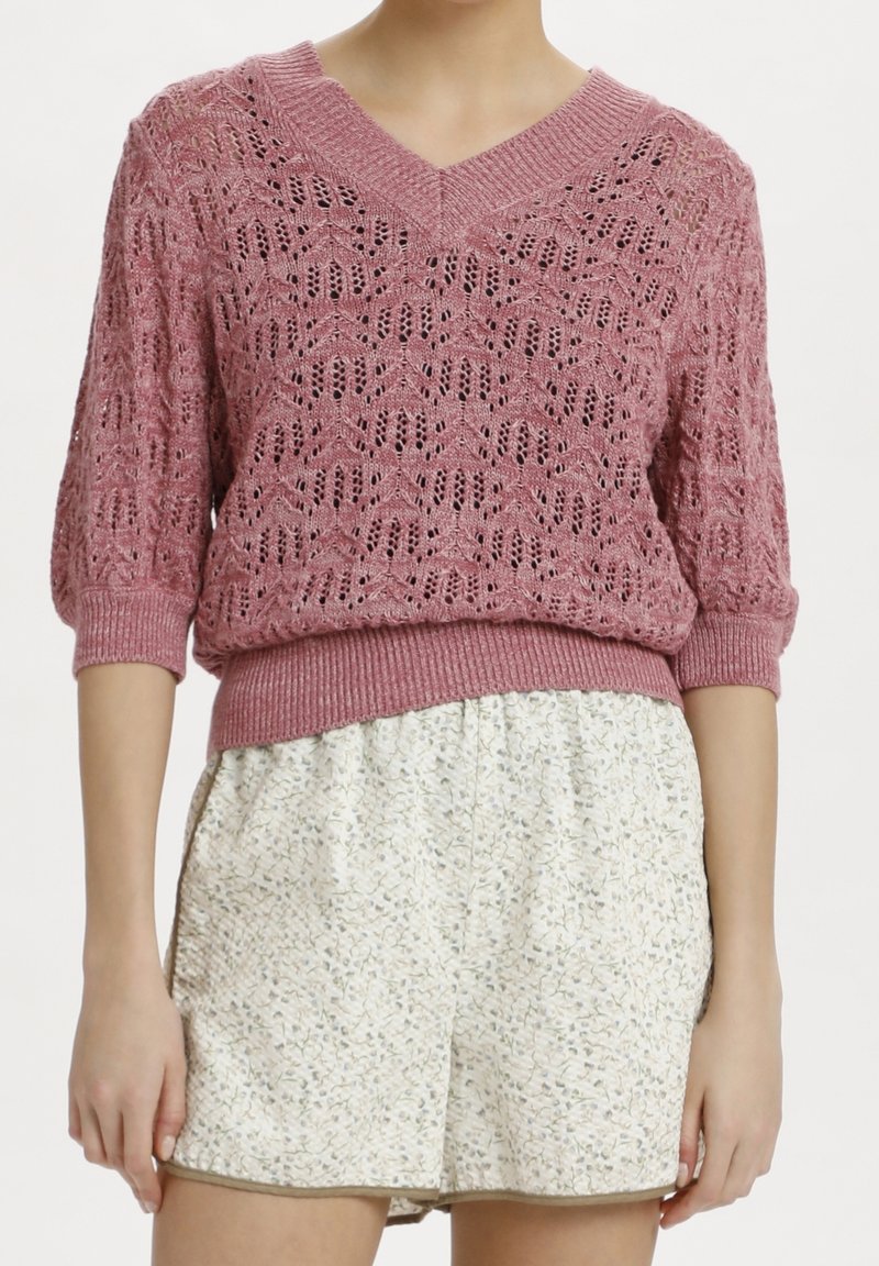 Femme portant un pull rose en maille à col en V avec des manches trois-quarts et un short blanc taille haute à motif floral.