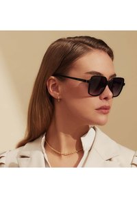 Lunettes de soleil noires avec des verres sombres, montures en métal fines et accents dorés. Portées avec une veste légère et un collier à chaînes dorées superposées.