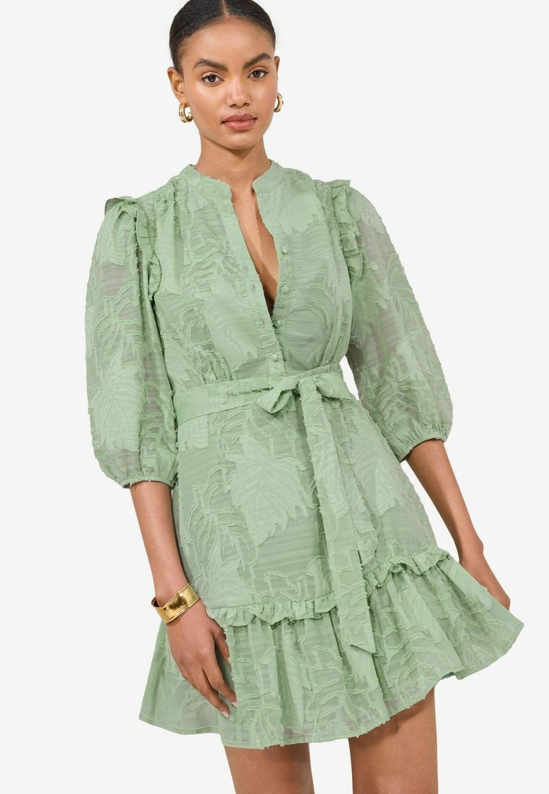 Hellgrünes Kleid mit Kragen, strukturiertem Blattmuster, puffärmel, Knopfleiste, tailliert und mit Rüschensaum. Goldene Armreif.