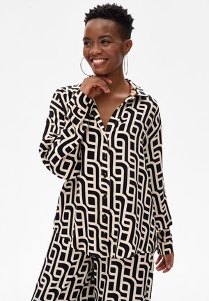 Uśmiechnięta kobieta w koszuli zapinanej na guziki z geometrycznym wzorem w kolorach czarnym i beżowym oraz dopasowanych spodniach, z dużymi kolczykami kołami.