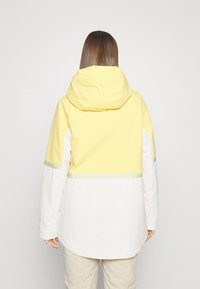 Gelbe und weiße Jacke mit Kapuze, ausgestattet mit einem horizontalen Farbblockdesign, reflektierenden Akzenten und einem lässigen Schnitt. Glatte Textur.