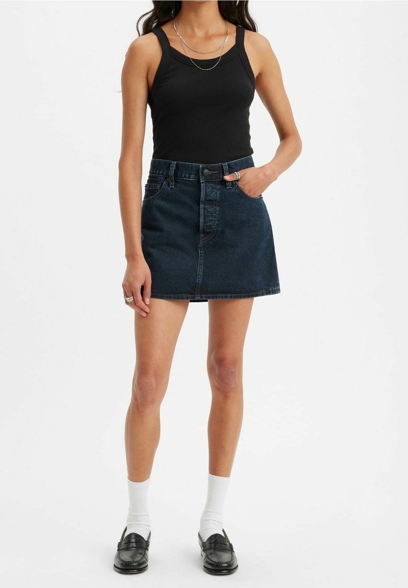 Femme portant un débardeur noir, une jupe en jean bleu foncé, des chaussettes blanches et des mocassins noirs, debout avec une main dans la poche.