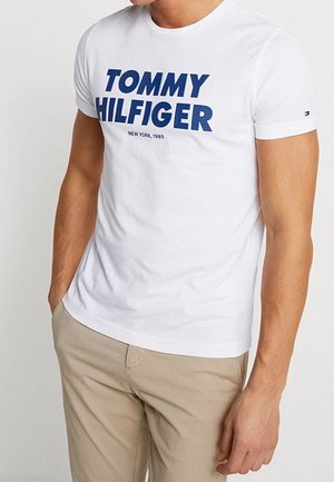 Mies, jolla on päällään valkoinen Tommy Hilfiger -t-paita sinisellä tekstillä ja beiget housut, seisoo käsi yhdessä taskussa.