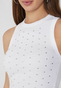 Top sin mangas de color blanco con textura acanalada y tachuelas plateadas cuadradas espaciadas uniformemente en la parte frontal. Textura suave y escote redondo.