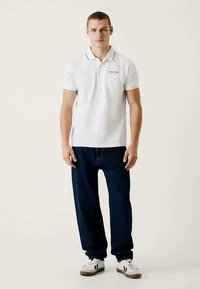 Ung mann står, iført hvit polo skjorte, mørkeblå jeans og hvite sneakers, mot en enkel lys bakgrunn.