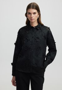 Zwarte button-up shirt met bloemmotieven en textuur; heeft een klassieke kraag en lange mouwen, wat een gestructureerde silhoët creëert.