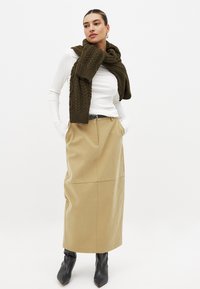 Long beige skirt in a smooth fabric, paired with a white ribbed long-sleeve shirt and a textured olive green knit scarf. Black pointed ankle boots.