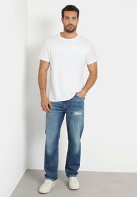 Tricou alb din bumbac cu logo-ul embosat "GUESS", asociat cu blugi bleu deschis, cu uzură subtilă, completat de adidași albi.