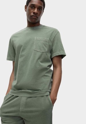 Mann trägt hellgrünes T-Shirt mit kurzen Ärmeln und Brusttasche sowie passende Hose, steht mit den Händen in den Taschen vor einem einfarbigen Hintergrund.