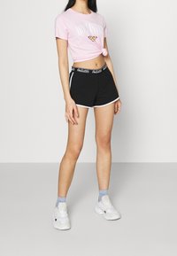 Růžové zkrácené tričko s logem Hollister, spárováno s černými sportovními šortkami s bílým lemem a elastickým pasem s logem. Bílé tenisky dotvářejí celý vzhled.