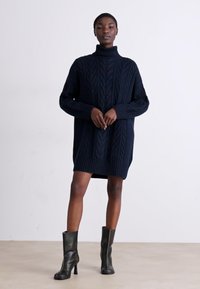 Σκούρο ναυτικό oversized φόρεμα πλεκτής ύφανσης με ψηλό στυλ ζιβά γυρισμένο και ρίγες στα μανίκια, συνδυασμένο με μαύρες μπότες με τακούνι μέχρι το γόνατο.