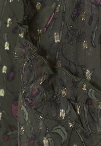 Mörkgrön tyg med blommönster i rosa och guldaccent, med volangkanter och metalliska utsmyckningar genom hela. 