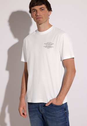 Mand iført hvid T-shirt med sort tekst på brystet står mod hvid baggrund med skygge på væggen.