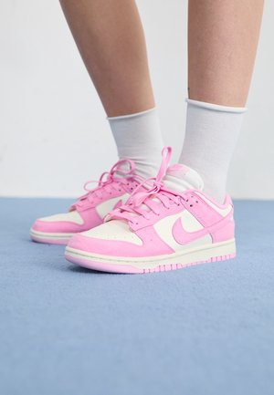 Osoba w białych skarpetkach i różowo-białych butach Nike stoi na jasnoniebieskiej podłodze przy białej ścianie.
