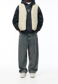 Kremowa pikowana kamizelka na ciemnej bluzie z kapturem, zestawiona z jasnoniebieskimi dżinsami. Białe trampki dopełniają casualowy wygląd.