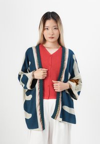 Vêtement surdimensionné multicolore avec des formes abstraites ; bleu marine, beige et accents. Présente un devant ouvert et des manches larges. Pull rouge en dessous.