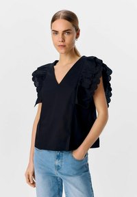Svart kortärmad topp med scallop-formade, lager-på-lager tygdetaljer vid axlarna, V-ringning och lös passform. Bärs med ljusblå jeans.