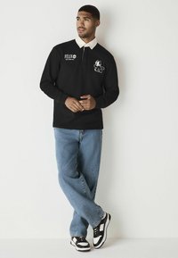 Schwarz langärmliges Poloshirt mit einem weißen Kragen, das einen Cartoon-Katzen-Druck und ein Logo auf der Brust zeigt. Kombiniert mit hellblauen Jeans und schwarzen Sneakers.