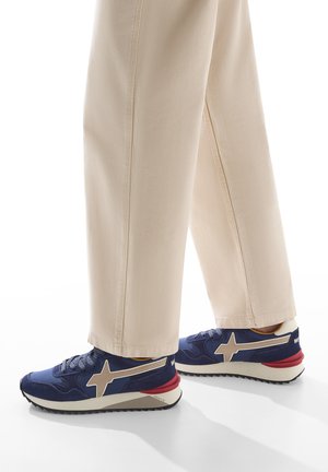 Sneaker navy con tomaia in tessuto texture, accenti in stelle beige, suola in gomma rossa e lacci blu, abbinati a pantaloni ampi color crema.