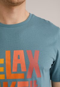 Türkiz pamut póló kerek nyakkivágással, elöl merész, több színű "RELAX" grafika található narancssárga, sárga és piros színekben.