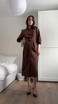 Γυναίκα που στέκεται με καφέ μίντι φόρεμα με ζώνη, μαύρα τακούνια, πόζα σε ένα μίνιμαλ υπνοδωμάτιο με λευκούς τοίχους και ξύλινο δάπεδο.