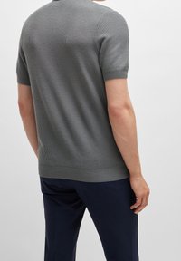Homme portant un pull en tricot gris à manches courtes et un pantalon bleu foncé, vu de dos contre un fond clair.
