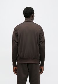 Καφέ αθλητικό σακάκι με ψηλό γιακά, ριγέ μανσέτες και μέση, κατασκευασμένο από λεία, ελαφριά ύλη, χωρίς εμφανή λογότυπα ή σχέδια.