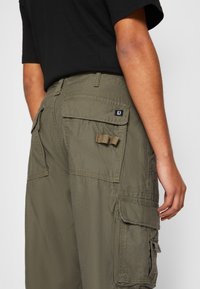 Khaki cargo kalhoty s volným střihem, které mají více bočních kapes, prošití a zadní nášivku s logem. Materiál se zdá být lehký.