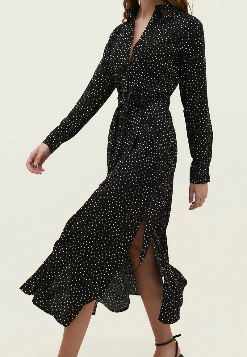 Robe midi noire à manches longues avec pois blancs, taille ceinturée et fente avant dévoilant la jambe du mannequin et des talons noirs à bride à la cheville.
