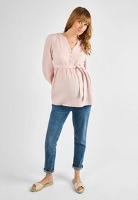Top lungo a maniche lunghe color rosa chiaro con scollo a V, dettagli con bottoni e vita annodata. Abbinato a jeans blu arrotolati e sandali beige con nodo.