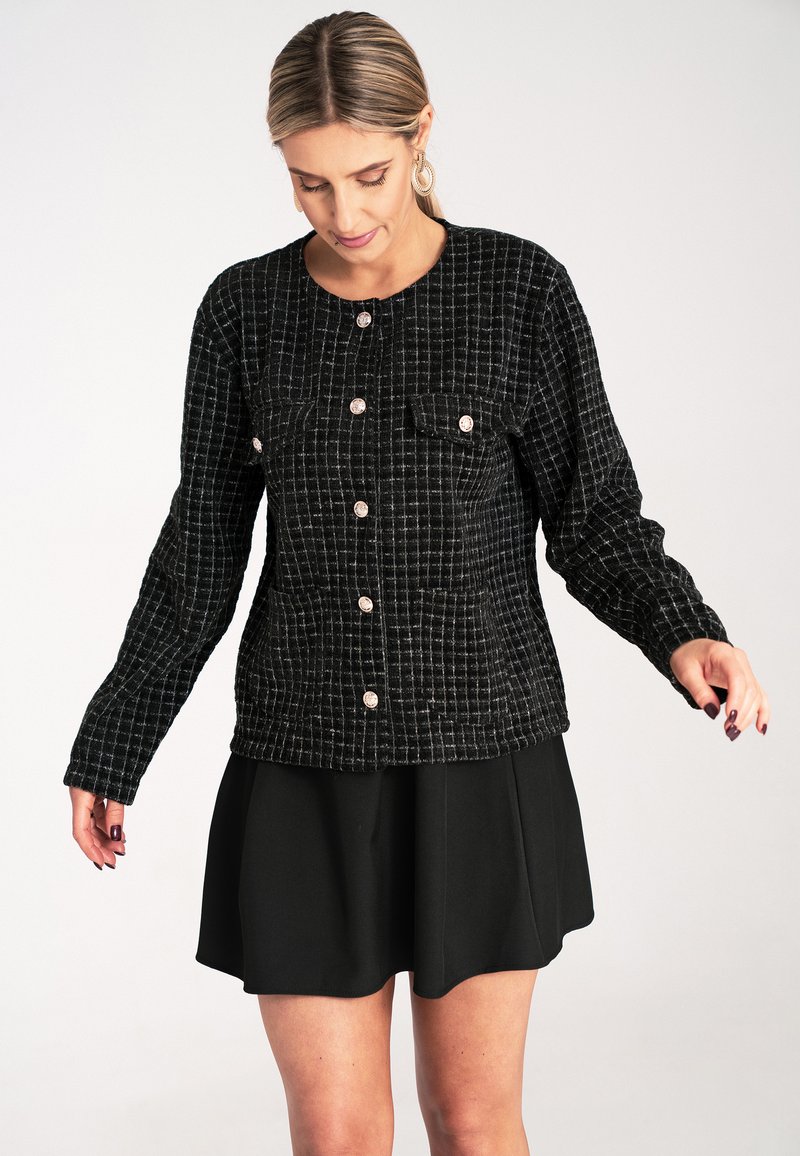 Czarnomartońska kurtka w kratę z długimi rękawami, okrągłym dekoltem, kieszeniami z przodu i metalowymi zapięciami, zestawiona z czarną spódnicą.