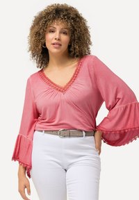 Blusa rosa con scollatura a V, bordo in pizzo e maniche a campana; abbinata a pantaloni bianchi e una cintura grigio chiaro. Tessuto morbido.