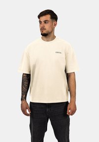 Krémové oversized tričko vyrobené z měkké látky, s kulatým výstřihem a bočním rozparkem. Černé logo "CRYSTAL" na levé části hrudi.