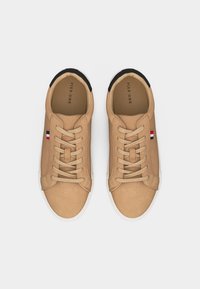 Ζευγάρι μπεζ αθλητικά παπούτσια με κορδόνια, λευκές σόλες και μικρή λεπτομέρεια με κόκκινη, λευκή και μπλε σημαία στις πλευρές, σε λευκό φόντο.