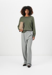 Maglione oversize verde oliva, pantaloni grigi chiari plissettati, in mano una borsa a tracolla beige, ballerine nere con punta a punta, ambiente interno.