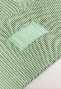 Heleheleroheline sooniline kangas tekstuuriga pinnaga. Lisandub õmmeldud ristkülikukujuline plaastriga kirjaga "self." samas toonis.