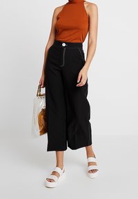 Pantalones negros de pierna ancha con costuras blancas y botón delantero, combinados con un top de halter acanalado naranja y sandalias de plataforma blancas. También se incluye un bolso transparente.