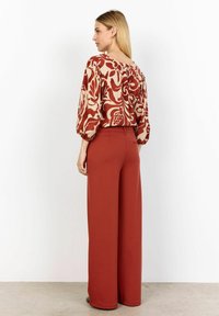 Bluza boje krem s cvjetnim uzorkom i napuhanih rukavima u kombinaciji s hlačama širokog kroja u boji rđe. Glatka tekstura i opušteni kroj.