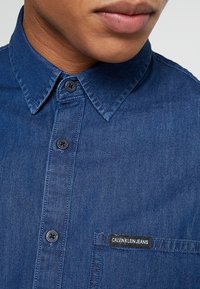 Mörkblå denimskjorta med klassisk krage, knäppning framtill och en bröstficka med en svart etikett som står "CALVIN KLEIN JEANS."