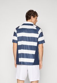 Mand, der står med ryggen til, iført en marineblå og hvid vandretstribet kortærmet skjorte og hvide shorts på en ensfarvet baggrund.