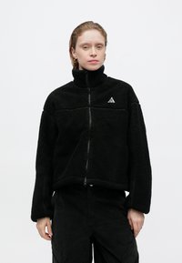 Geacă din fleece neagră cu guler înalt, fermoar frontal și suprafață texturată; are un mic logo triunghiular alb pe piept.