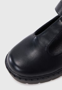 Black leather shoe featuring a rounded toe, strap across the top, and cutout detail. Textured sole with ridges for traction.