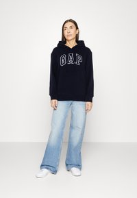 Navy blue hoodie with "GAP" logo, drawstring hood, long sleeves, and front pocket; paired with light blue flared jeans and white sneakers.