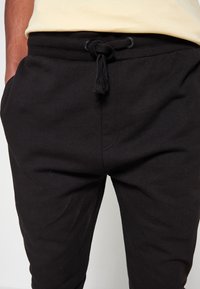 Un pantalon de survêtement noir en tissu mélangé de coton, doté d'une taille élastique avec cordon de serrage et de poches latérales. Texture lisse et mate.