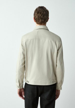 Beige Denimjacke mit lockerem Schnitt, spitzem Kragen und geknöpften Manschetten. Mit Rückennaht und verstellbaren Seitentaschen. Schwarze Jeans sind darunter sichtbar.