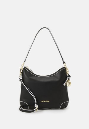 Schwarze Schultertasche aus synthetischem Material mit weißer Paspelierung, goldfarbenen Metallbeschlägen und einem Anhänger. Sie hat ein geschwungenes Design und einen verstellbaren Riemen.