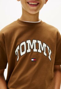 Bruine katoenen T-shirt met witte "TOMMY" letters en een klein rood, wit en blauw logo accent. Korte mouwen, ronde hals ontwerp.